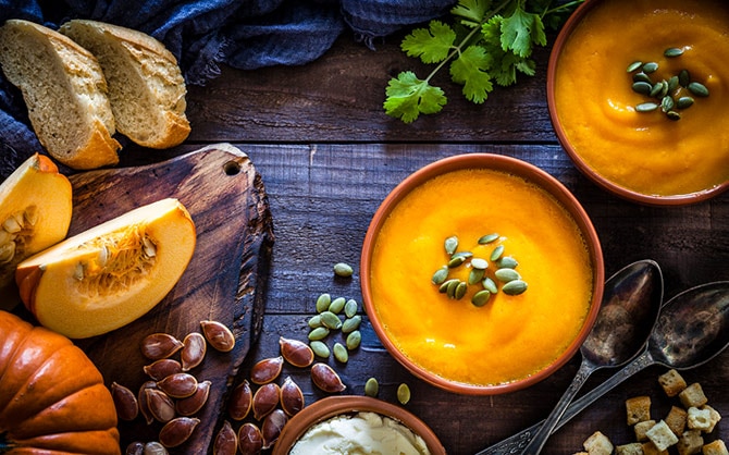 Una imagen de dos bol de sopa de calabaza con pipas de calabaza y rodeados de rebanadas de pan rústico, un par de cucharas, unos tropiezos de pan, una ramita de cilantro y una calabaza entera.