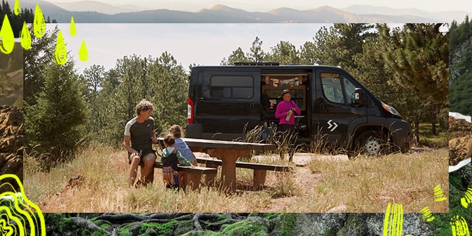 Ein Mann sitzt mit zwei Kindern an einem Picknicktisch, während eine Frau neben dem Camper steht